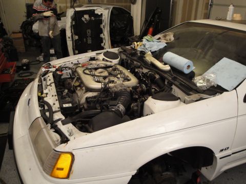 1989 Ford Taurus - The engine bay of a white car, identified by a '3.0' badge, is open for work, with another vehicle visible in the background.