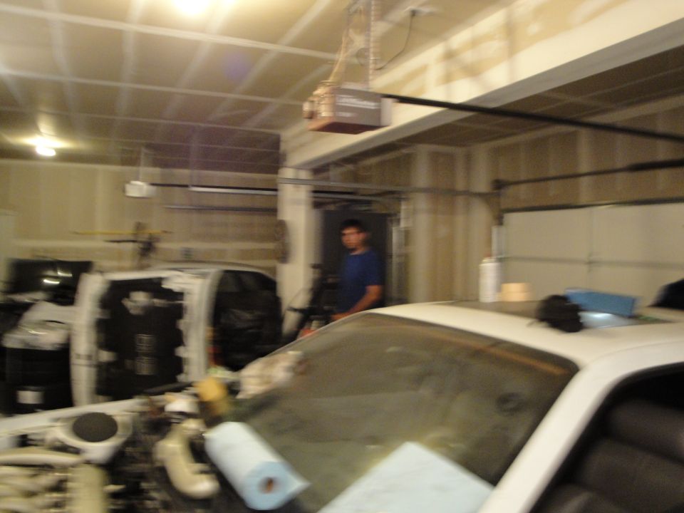 White car interior and engine bay with tools and paper towels in a garage. A person stands in the background