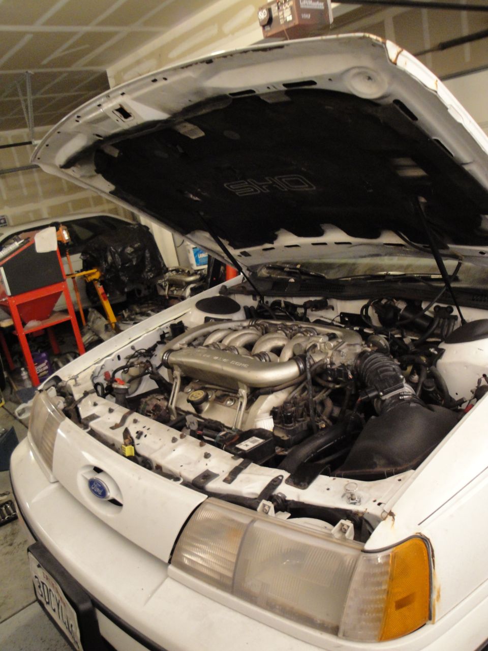 White Ford Taurus SHO with hood open, showing V6 engine. Another car is partially visible in the background