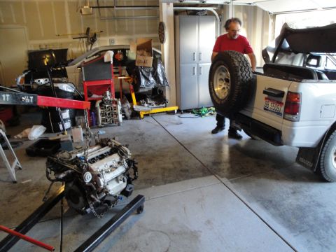 1989 Ford Taurus - A man loads a spare tire onto a white Isuzu pickup truck in a garage, with a detached engine on a stand nearby.