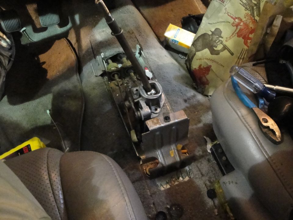 Car interior showing a disassembled transmission shifter mechanism, tools, and a bag with graphics