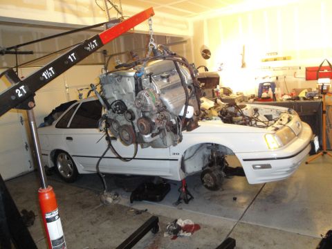1989 Ford Taurus - A car's engine is suspended by a hoist, ready for removal or installation, with the vehicle partially disassembled.