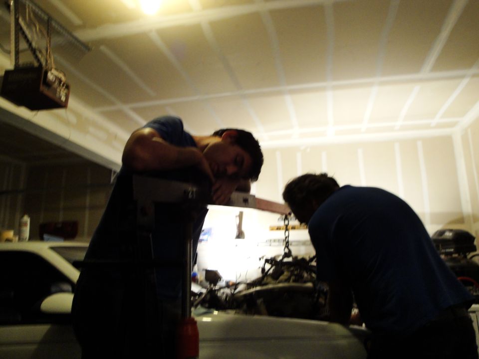 Two people working on an engine in a garage with an engine hoist