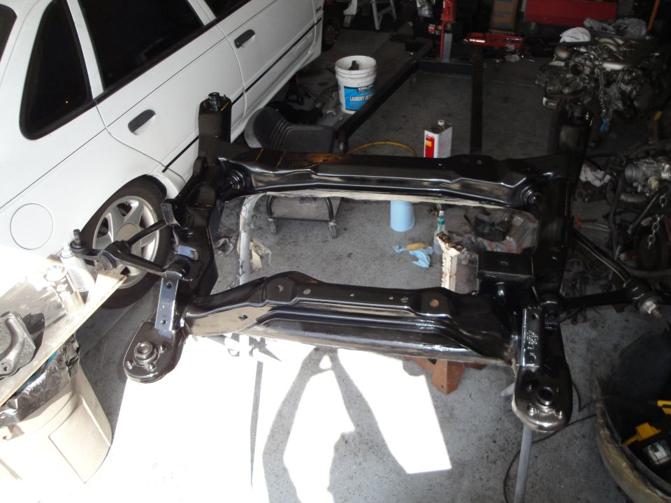 Black painted subframe assembly with suspension components and a white car in the background