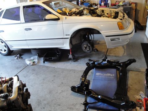 1989 Ford Taurus - A white sedan is in a garage with its front end disassembled and parts laid out on the floor.