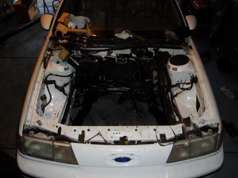 1989 Ford Taurus - The engine bay of a white 1989 Ford Taurus shows the empty space where the engine was removed, indicating an engine swap is in progress.