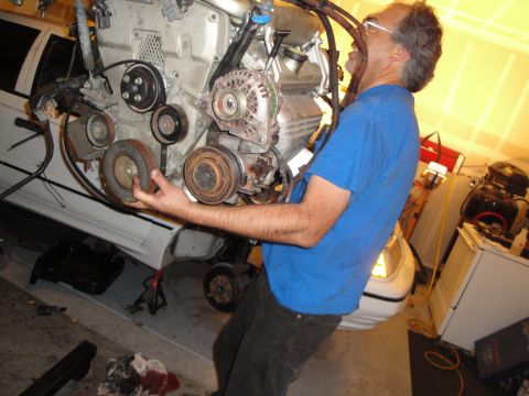 1989 Ford Taurus - A man is shown holding a car engine, with a white vehicle partially visible in the background, suggesting an engine swap or repair.