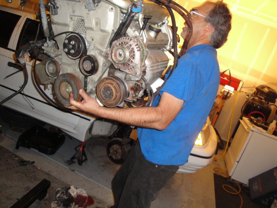 Man holding a car engine, with a white car in the background