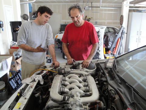 1989 Ford Taurus - Two men are gathered around a car engine, appearing to be in the process of working on it within a garage setting.
