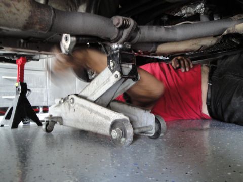 1989 Ford Taurus - Underneath a vehicle, a mechanic works on the exhaust system using a floor jack and jack stands.