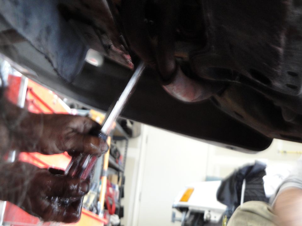 Hands in oil working on car exhaust pipe, another car visible in background