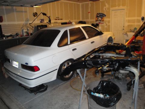 1989 Ford Taurus - A white 1989 Ford Taurus sedan is parked in a garage, with engine components laid out in front of it.