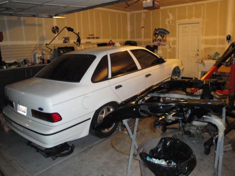 1989 Ford Taurus - A white sedan is parked in a garage, with a disassembled black bumper assembly in front of it.