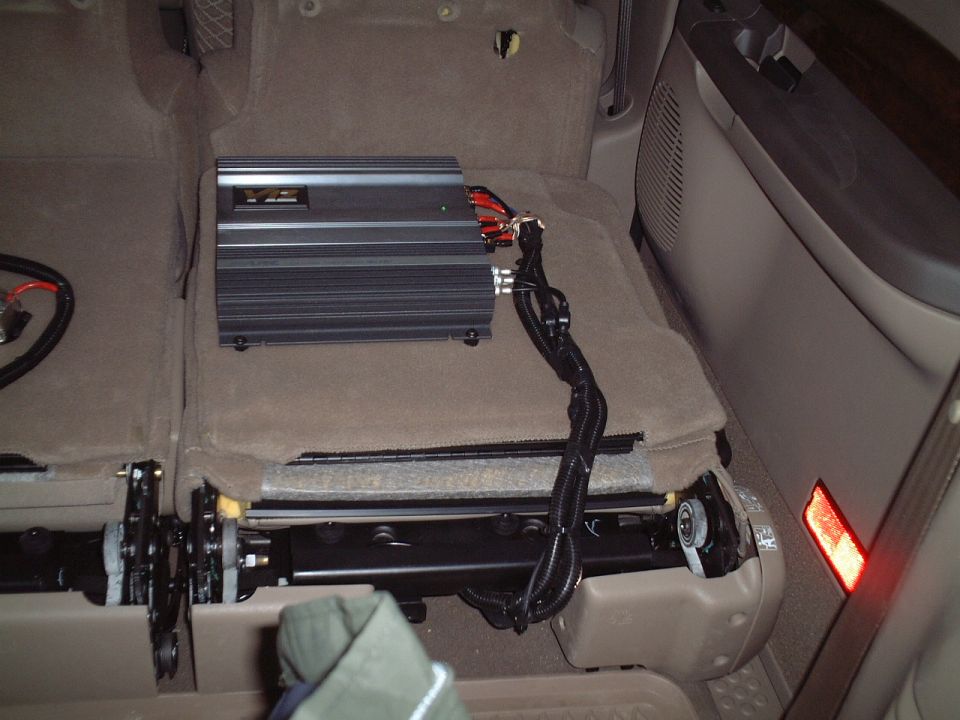Black V12 amplifier mounted on tan carpet under rear seat area with wiring harness.