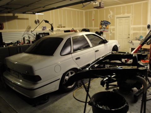 1989 Ford Taurus - A white sedan is parked in a garage, with parts of another vehicle in the foreground.