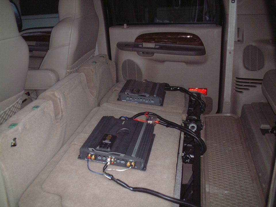 Two black car amps mounted on tan folded seat in SUV rear with power and RCA wiring
