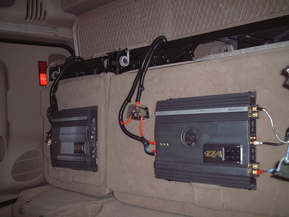 Two black car audio amplifiers with wiring mounted on tan carpet behind SUV rear seats.