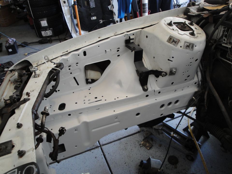 White car engine bay with exposed metal and wiring harnesses. Some parts are removed