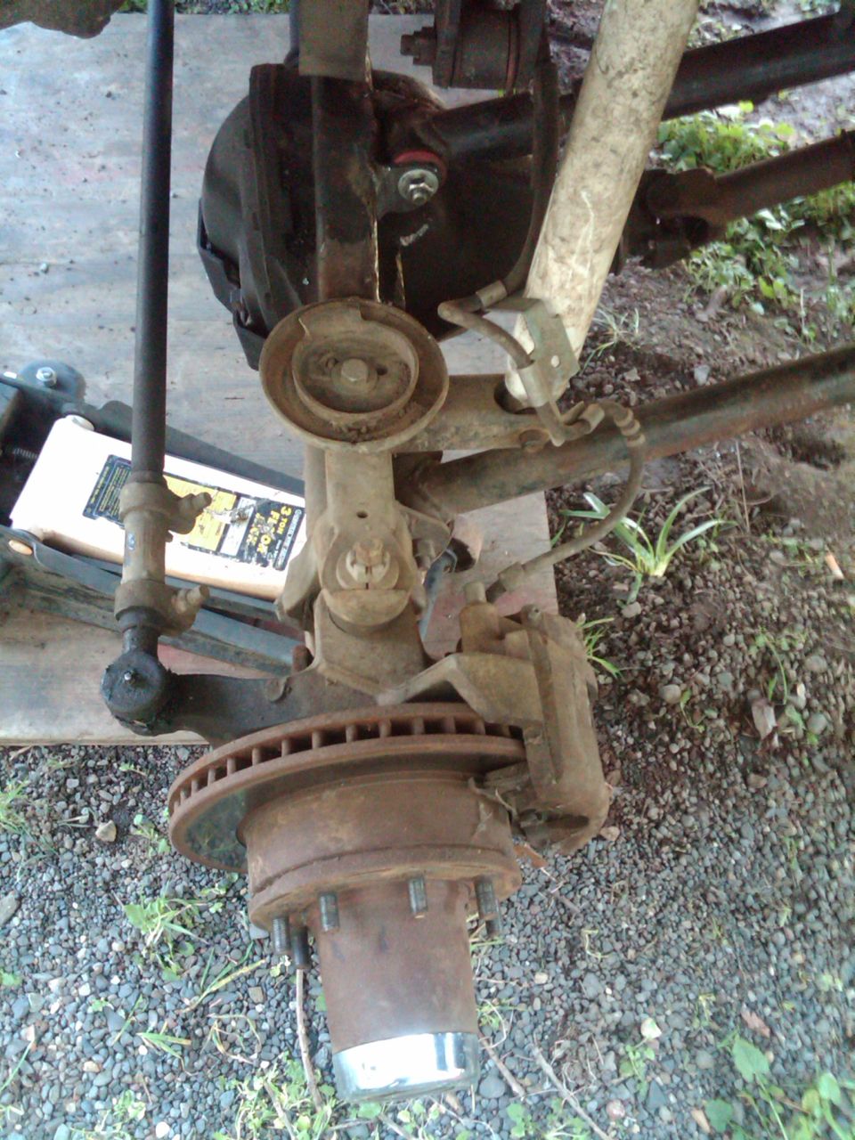 Rusty front solid axle knuckle, rotor, hub and radius arm removed and lying on gravel