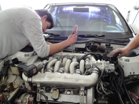 1989 Ford Taurus - Mechanics work on the engine of a car, focusing on the intake manifold and various hoses.
