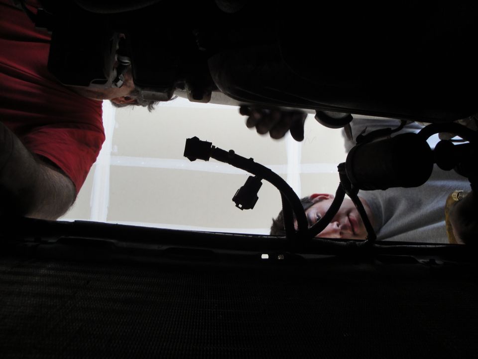 Underside view of an engine bay with two people working, showing wiring and mechanical parts
