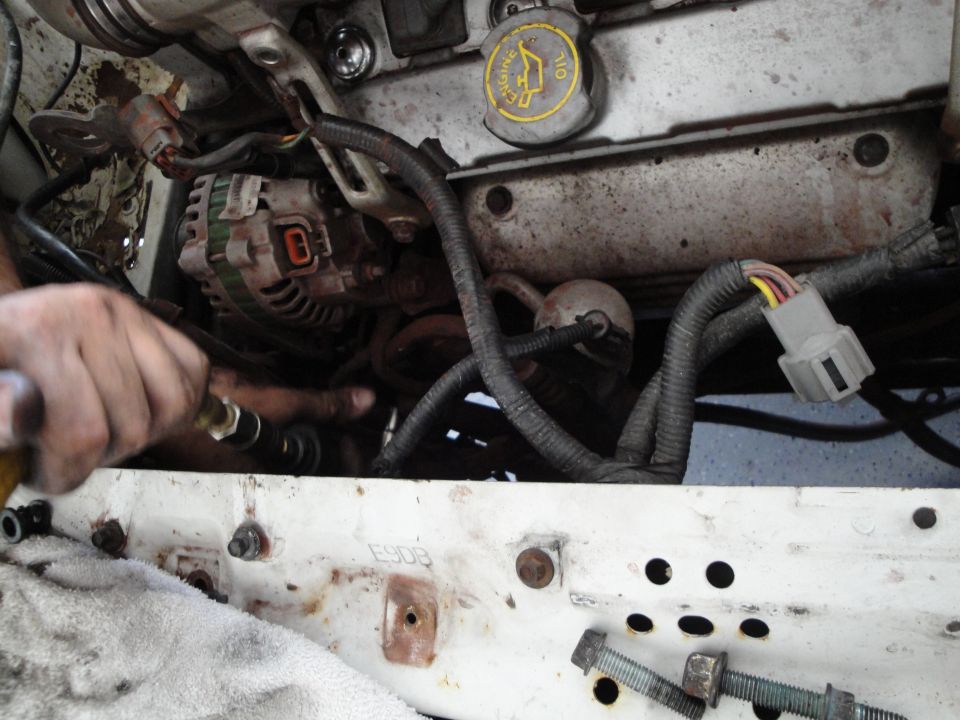 Engine bay with alternator, wiring, and an oil cap with Ford logo. A hand is working on a hose