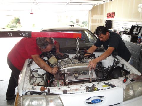 1989 Ford Taurus - Two mechanics work on an engine swap in the bay of a 1989 Ford Taurus, using an engine hoist.