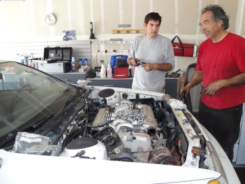 1989 Ford Taurus - Two men are gathered around the open engine bay of a white car, appearing to be in the middle of a repair or modification.