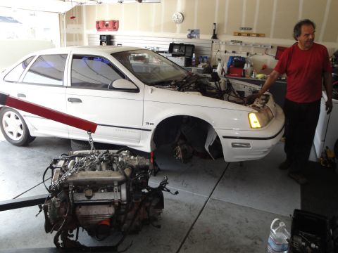1989 Ford Taurus - A man stands by a white sedan in a garage as its engine is suspended by a hoist.