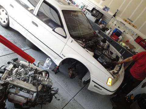 1989 Ford Taurus - A white 1989 Ford Taurus is shown in a garage with its engine removed and suspended by a hoist, indicating an engine swap or repair.