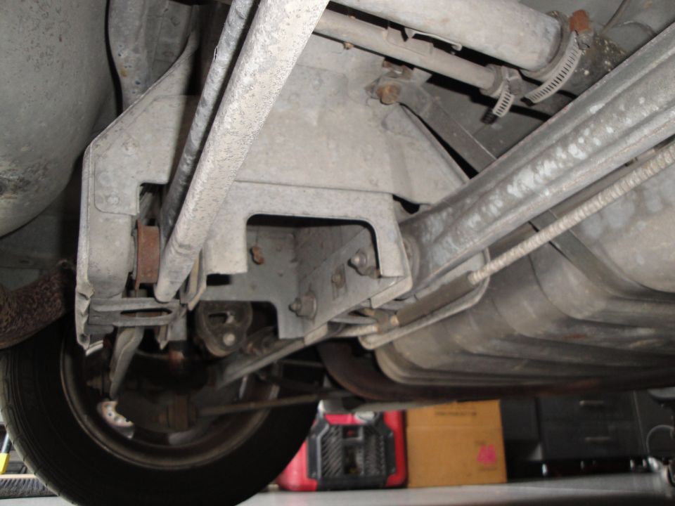 Underside view of a car's rear suspension, showing control arms, a fuel tank, and a tire