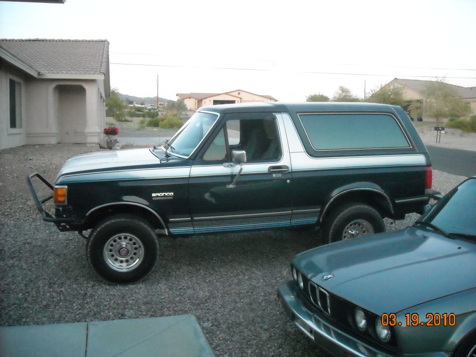 1988 Ford Bronco, dark green with silver top, aftermarket front bumper. Another car is partially visible in the foreground