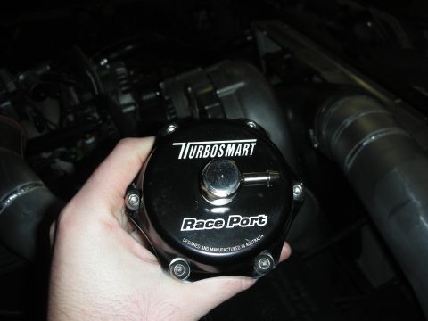 1992 Ford Crown Victoria - A Turbosmart Race Port blow-off valve is held in front of a vehicle's engine bay.
