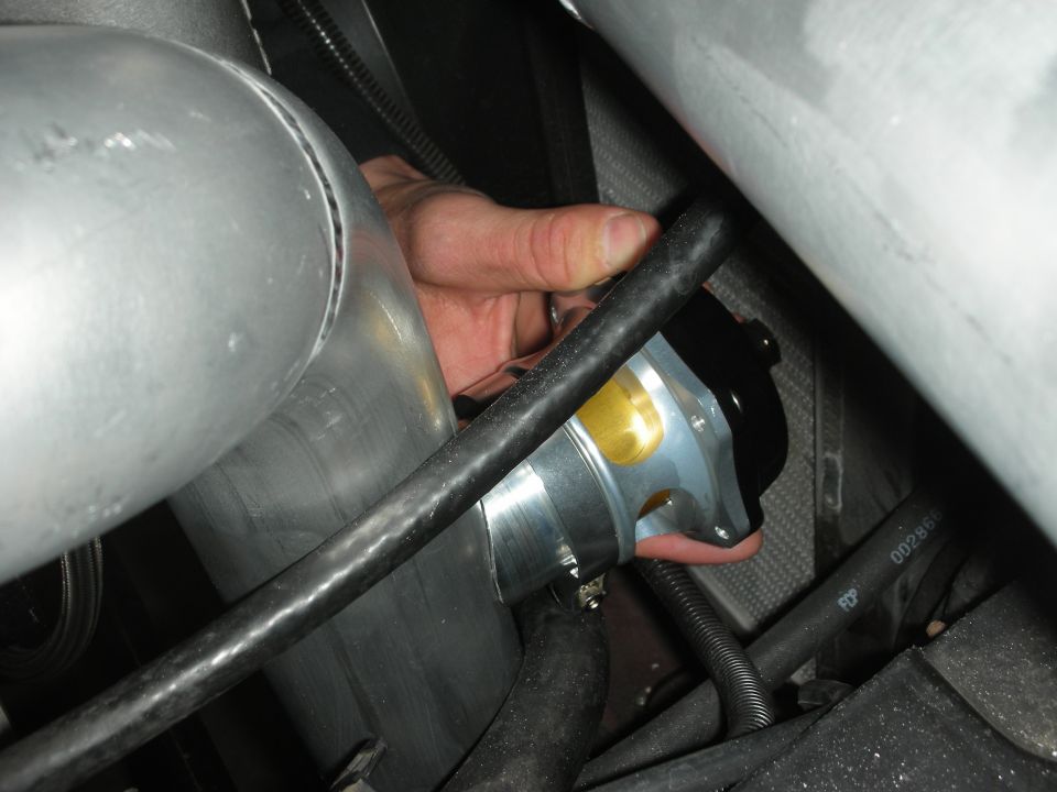 Close-up of a hand installing a blow-off valve on a large aluminum pipe in an engine bay.