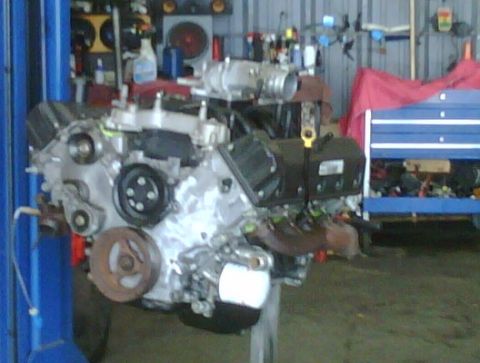 2005 Ford Crown Victoria - Engine assembly, likely a V8, is suspended from a hoist in a garage setting with tools and equipment visible in the background.