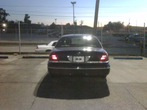 2005 Ford Crown Victoria - Rear view of a dark 2005 Ford Crown Victoria parked in a lot at dusk. A white vehicle is visible behind a fence in the background.