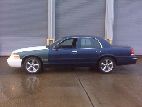 2005 Ford Crown Victoria - A blue and white sedan with aftermarket wheels is parked in front of a large garage door.