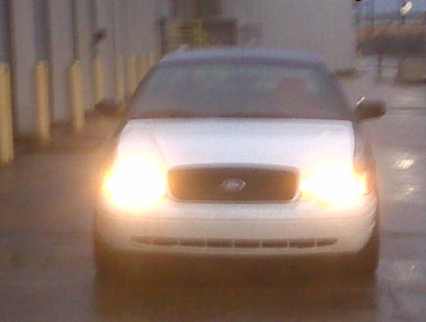 2005 Ford Crown Victoria - The front of a white Ford Crown Victoria is visible with its headlights illuminated.