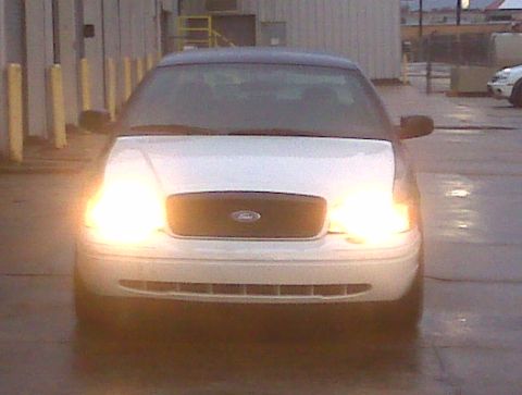 2005 Ford Crown Victoria - A white 2005 Ford Crown Victoria is parked outside on a wet day, with its headlights illuminated.