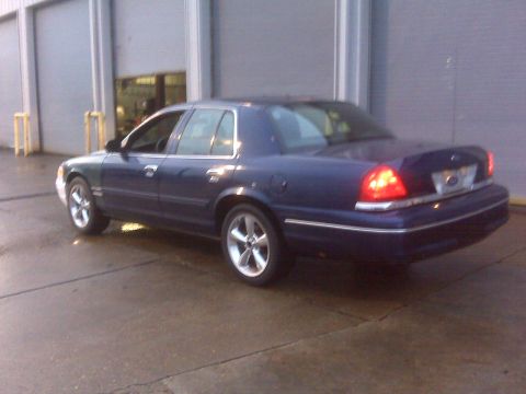 2005 Ford Crown Victoria - A dark blue 2005 Ford Crown Victoria is seen from a three-quarter front angle, parked on a wet surface in front of a garage door.