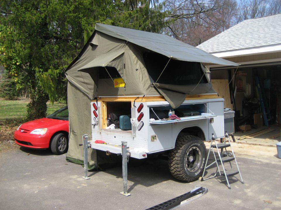 Off-road trailer with rooftop tent and a red sedan parked nearby