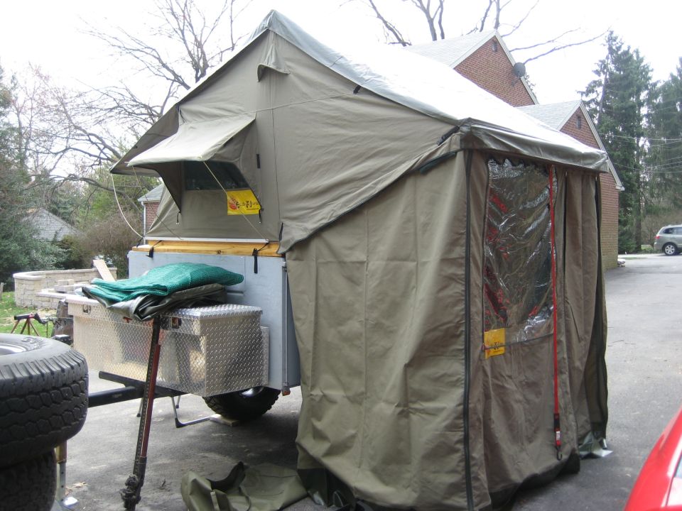 Pop-up camper trailer with attached tent and storage boxes. Spare tire visible on the left