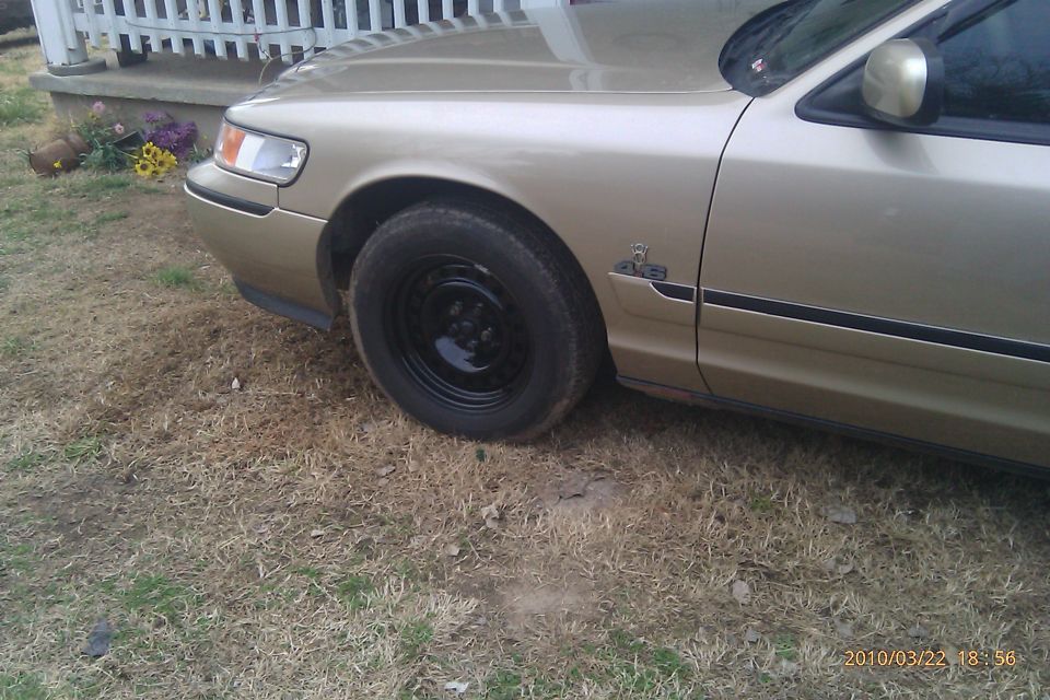 Gold sedan, possibly a Ford Crown Victoria or Mercury Grand Marquis, parked on dry grass. Shows front fender, door, and wheel