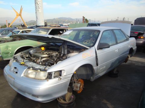 1993 Ford Taurus - This silver sedan, identified as a 1993 Ford Taurus, is in a junkyard with its hood open and missing wheels.