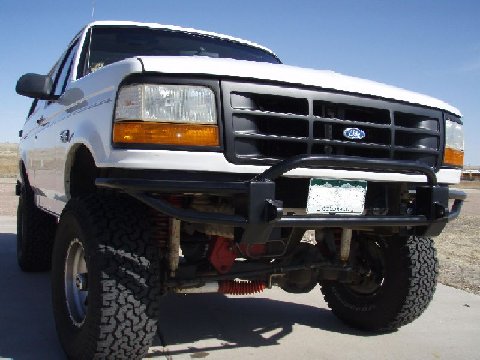 1993 Ford Bronco - Front three-quarter view of a lifted white Ford Bronco outdoors on gravel, wearing a black tube front bumper with brush-guard uprights and…