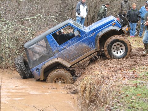 1985 Ford Bronco - A blue off-road vehicle is stuck in a muddy stream while onlookers observe the situation.