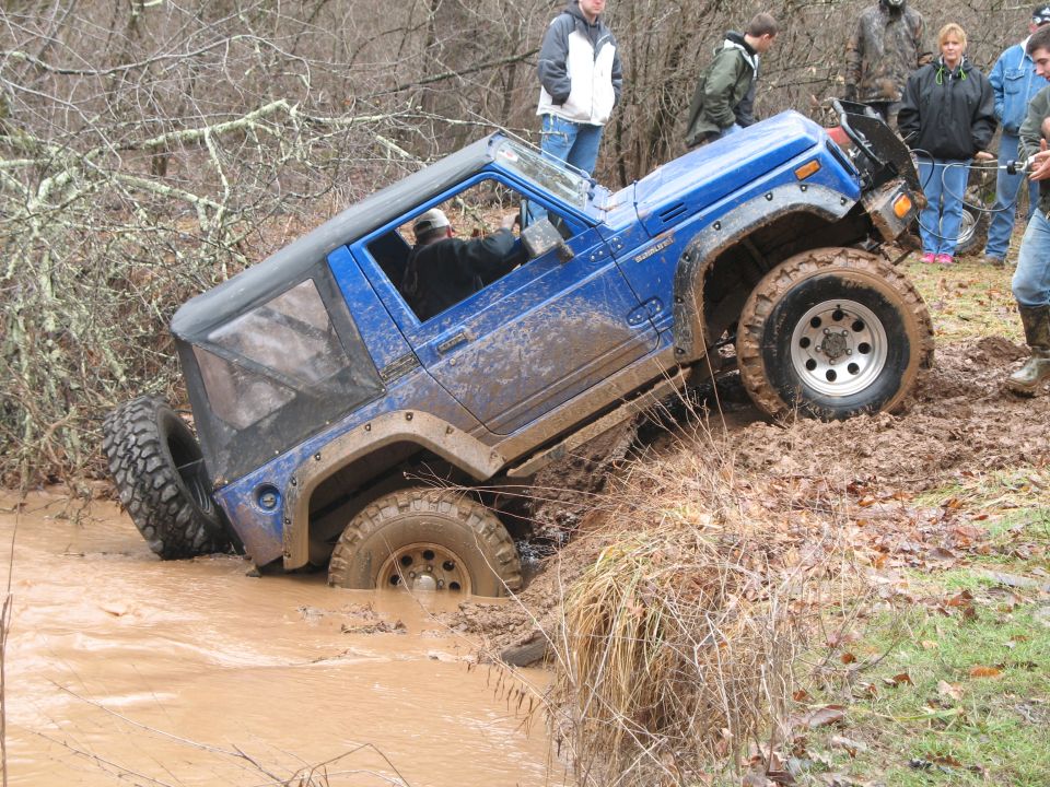 Mud-covered blue off-road vehicle tilted in a muddy ditch with people watching