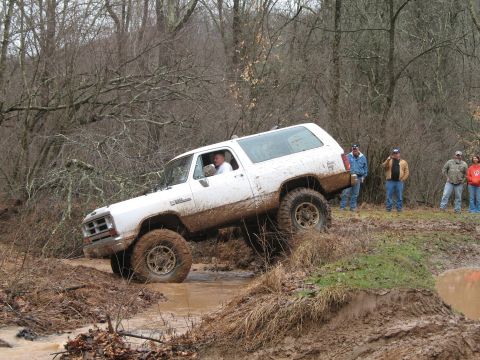 1985 Ford Bronco - White off-road SUV navigates a challenging muddy terrain, with spectators observing the action.