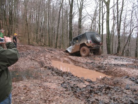 1985 Ford Bronco - A Jeep Wrangler is stuck in a muddy trail, with onlookers watching.