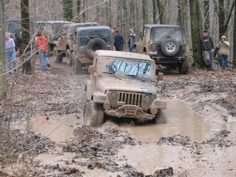 1985 Ford Bronco - Several muddy off-road vehicles navigate a challenging course through a wooded area.